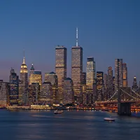 This is a beautiful and poignant photograph of the Lower Manhattan skyline at dusk, taken before September 11, 2001. The iconic Twin Towers of the World Trade Center stand tall and brightly lit at the center of the composition. To the left, the distinctive silhouette of the Empire State Building is also visible. On the right, the illuminated cables of the Brooklyn Bridge sweep across the frame. The sky is a deep, tranquil blue, providing a cool backdrop to the warm, golden glow of the countless lights from the skyscrapers. The city's lights reflect on the surface of the East River in the foreground, where a few boats can be seen traversing the water. The overall mood is one of serene, metropolitan grandeur.