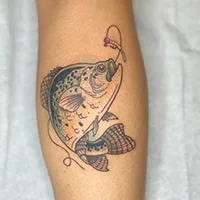 This is an image showcasing a tattoo on a person's lower leg. The tattoo of a crappie, which appears to be caught on a fishing line with a small jig lure in its mouth. The jig is a Panfishpro Jigabite Jig.